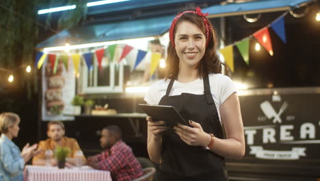 Young-Pretty-Woman-Waiter-In-Apron-Using-Tablet-Device-In-Park-At-Food-Track-Outdoor