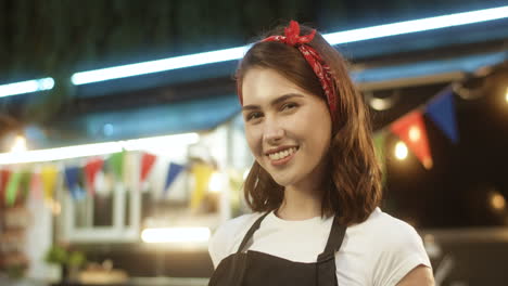 Portrait-Of-Attractive-Joyful-Young-Girl-In-Apron-Smiling-Happily-To-Camera-Outdoor-With-Small-Food-Truck-On-Background