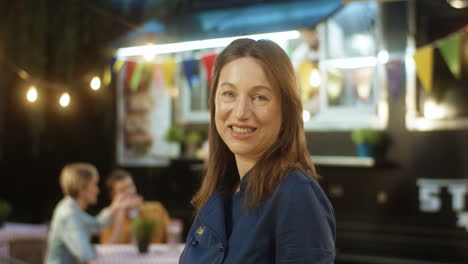 Close-Up-Of-Beautiful-Middle-Aged-Woman-In-Apron-Smiling-Happily-To-Camera-Outdoors-With-Little-Food-Truck-Bar-On-Background