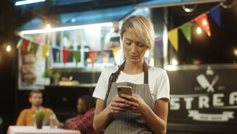 Young-Blonde-Woman-With-Short-Hair-In-Hair-Taping-And-Texting-Message-On-Smartphone-At-Festive-Food-Track-At-Night