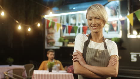 Happy-Beautiful-Joyful-Young-Woman-In-Apron-With-Blonde-Short-Hair-Smiling-At-Festive-Food-Track-In-Evening