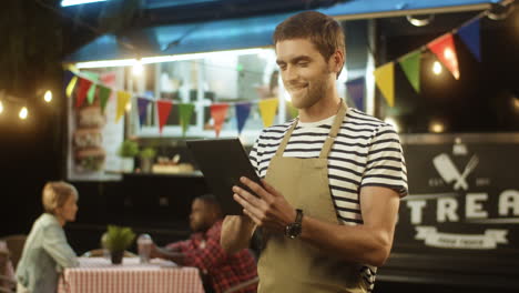 Young-Man-Waiter-In-Apron-Smiling-And-Using-Tablet-Device-In-Evening-At-Food-Truck-In-Festive-Park
