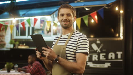 Portrait-Of-Young-Man-Waiter-In-Apron-Smiling-To-Camera-And-Using-Tablet-Device-In-Evening-At-Food-Truck-In-Festive-Park