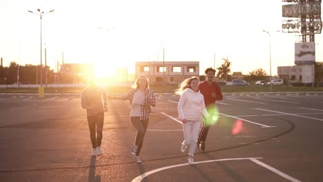 Group-Of-Four-Stylish-Friends-Are-Running-By-Empty-Parking-Zone-Outdoors-Fun-Happiness,-Young-Men-And-Women-Are-Running-In-The-Evening-Dusk-Then-Freerly-Jumping-Together