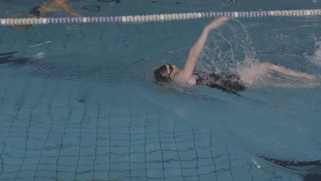Lateral-Tracking-Shot-Of-A-Young-Female-Swimmer-Swimming-Backstroke-In-An-Indoor-Pool