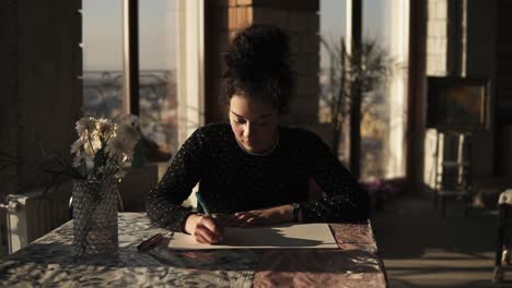 Young-Sophisticated-Female-Artist-Drawing-With-A-Pen,-Siting-By-The-Table-In-Art-Studio-With-Wide-Panoramic-Windows-On-The-Background