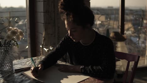 Young-Cute-Female-Artist-With-Curly-Hair-Pulled-In-A-Bun,-Sitting-By-The-Table-And-Drawing-With-A-Pen-Fully-Concentrated-On-The-Process-Of-Creation