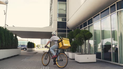 Food-Delivery-Courier-Wearing-Thermal-Backpack-Rides-A-Bike-Next-To-An-Office-Building-To-Deliver-Orders-For-Clients-And-Customers
