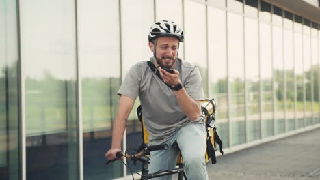 Happy-Food-Delivery-Man-Wearing-Thermal-Backpack-On-His-Bicycle-Sending-An-Audio-Message-With-His-Smartphone