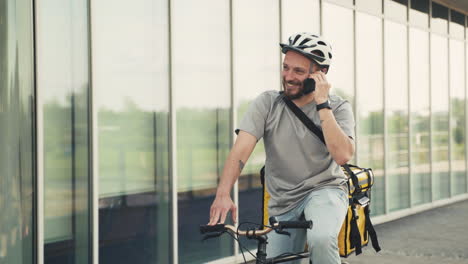 Happy-Food-Delivery-Man-Wearing-Thermal-Backpack-Having-A-Funny-Conversation-On-The-Phone-On-His-Bicycle