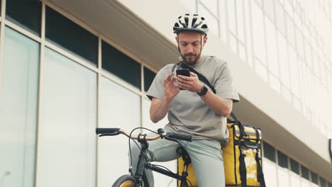 Food-Delivery-Guy-Wearing-Thermal-Backpack-Taking-A-Look-At-His-Smartphone-On-His-Bike