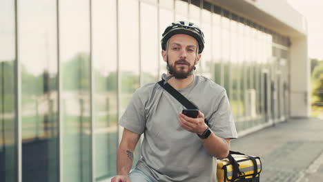 Confused-Food-Delivery-Guy-Wearing-Thermal-Backpack-Takes-A-Look-At-His-Smartphone-To-Know-Where-To-Go-With-His-Bike