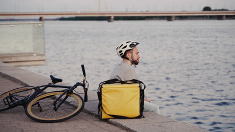Food-Delivery-Guy-Rests-Next-To-His-Thermal-Backpack-At-Sunset-On-The-Shore-Of-A-River-And-Takes-A-Look-At-His-Smartwatch-Waiting-For-The-Next-Order