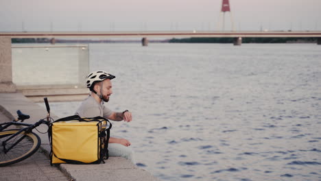Food-Delivery-Man-Rests-Next-To-His-Thermal-Backpack-At-Sunset-On-The-Shore-Of-A-River-And-Takes-A-Look-At-His-Smartwatch-To-Know-Where-To-Go-Next
