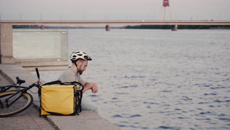 Food-Delivery-Man-Rests-Next-To-His-Thermal-Backpack-At-Sunset-On-The-Shore-Of-A-River-And-Consults-At-His-Smartwatch