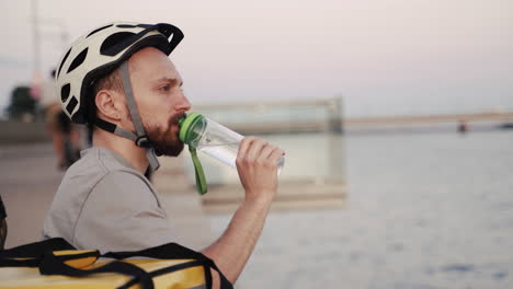 Food-Delivery-Guy-Seated-Next-To-A-River-At-Sunset-Drinks-Some-Water-During-His-Break