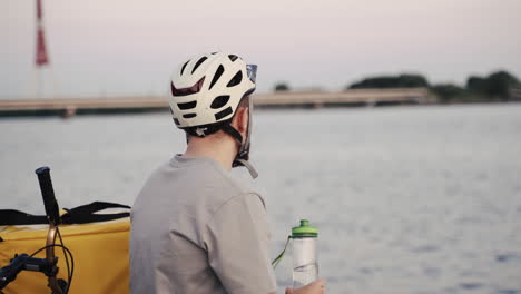 Exhausted-Food-Delivery-Man-Seated-Next-To-A-River-At-Sunset-Drinks-Some-Water-From-A-Plastic-Bottle-During-His-Break