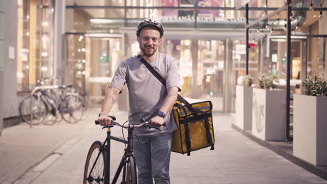 Happy-Bearded-Food-Delivery-Man-Wearing-A-Thermal-Backpack-And-Helmet-Looks-Directly-Into-The-Camera