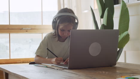 A-Pretty-And-Intelligent-Blonde-Girl-With-Headphones-Looks-At-The-Laptop-Screen-And-Does-Her-Homework-Using-A-Pencil-And-Paper