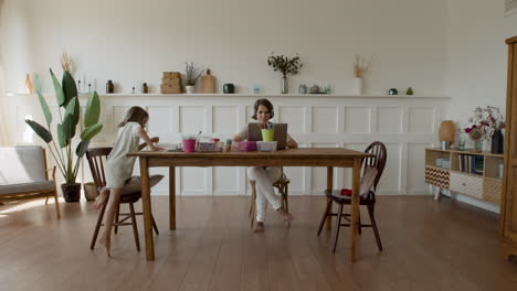 Wide-Shot-Of-A-Mother-Working-At-Home-Making-A-Videocall-When-Her-Daughter-Comes-In-And-Sits-Down-Next-To-Her-To-Do-Her-Homework