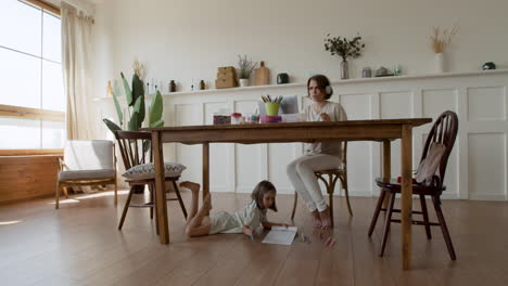 Wide-Shot-Of-A-Mother-Making-A-Videocall-While-Her-Daughter-Is-Distracted-Under-The-Table-Drawing-1