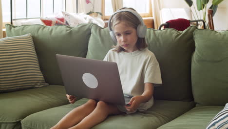 A-Little-Blonde-Girl-With-Headphones-Stares-Intently-At-The-Laptop-Screen-Seated-On-The-Sofa