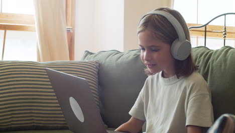 Close-Up-Of-A-Little-Blonde-Girl-With-Headphones-Having-Fun-While-Watching-At-The-Laptop-Screen-Seated-On-The-Sofa