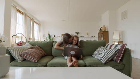 Wide-Shot-Of-A-Very-Affectionate-Mother-Who-Gives-Kisses-To-Her-Responsible-Little-Daughter-While-She-Does-Her-Homework-On-The-Laptop