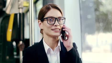 Close-Up-Of-The-Young-Pretty-Businesswoman-In-Glasses-Talking-On-The-Mobile-Phone-With-A-Smile-While-Sitting-In-The-Tram-And-Going-Home