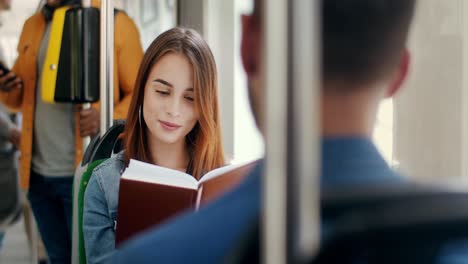 Young-Beautiful-And-Happy-Girl-Reading-A-Book-In-The-Tram-And-Smiling,-Then-Flirting-With-A-Young-Handsome-Man-Who-Sitting-In-Front-Of-Him