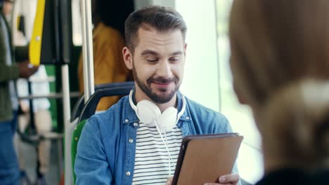 Young-Good-Looking-Happy-Man-With-Big-Headphones-On-His-Neck-Watching-Something-On-The-Tablet-Device-And-Laughing-Or-Having-Videochat-In-The-Tram