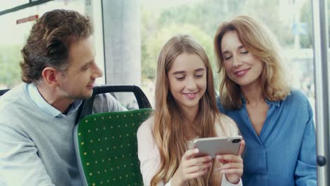 Pretty-Teen-Girl-Sitting-With-Her-Parents-In-The-Tram-And-Showing-Them-Something-Interesting-On-The-Smartphone