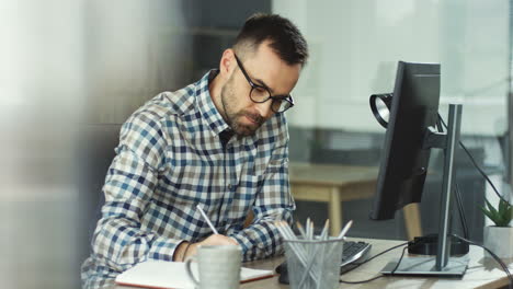 Young-Man-In-Glasses-Working-At-The-Table-In-His-Office-And-Writing-Down-Something-In-Front-Of-The-Computer-Screen