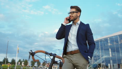 Attractive-Young-Man-In-Glasses-And-Bussiness-Style-Standing-With-A-Bike-Near-The-Big-Glass-Urban-Building-And-Talking-On-The-Mobile-Phone