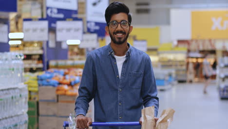 Portrait-Of-A-Tall-Boy-With-Glasses-Leaning-On-The-Shopping-Cart-In-The-Supermarket-Looking-Straight-At-The-Camera-And-Smiling