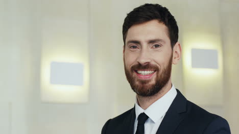 Portrait-Of-The-Young-Handsome-Businessman-With-Beard-And-In-A-Tie-Turning-His-Face-And-Looking-To-The-Camera-With-Smile-On-The-Wall-Background