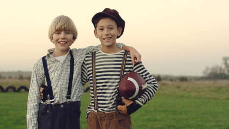 Portrait-Shot-Of-The-Two-Teen-Boys-Friends-Players-Of-American-Football-Standing-With-A-Ball-And-Embracing-Each-Other-On-The-Field-Of-The-Countryside,-Looking-At-Each-Other-With-Smiles-And-Then-To-The-Camera
