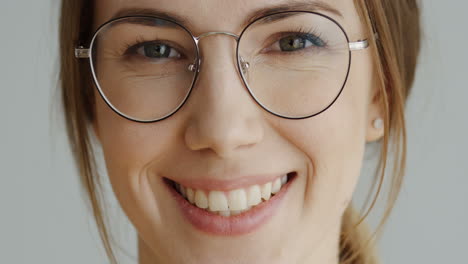 Close-Up-Of-The-Woman's-Face-In-Glasses-With-Fair-Hair-Looking-Straight-In-The-Camera-And-Smiling-On-The-White-Wall-Background