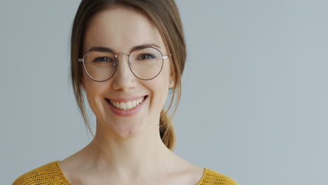 Portrait-Of-The-Beautiful-Woman-In-Glasses-And-With-Long-Fair-Hair-Looking-Straight-In-The-Camera-Seriously-And-Then-Smiling-Cheerfully-On-The-White-Wall-Background