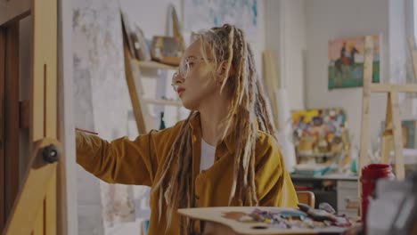 Close-Up-Of-Young-â€“Â°Auc-Beautiful-Girl-In-Glasses-And-Dreadlocks-Drawing-At-Easel-In-Cozy-Messy-Studio
