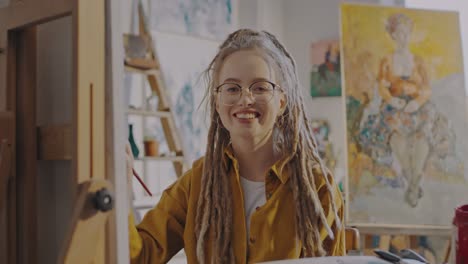 Portrait-Of-Young-Pretty-Female-Student-Of-Fine-Art-In-Glasses-And-With-Dreadlocks-Painting-At-Easel-In-Cozy-Workshop-And-Smiling-To-Camera
