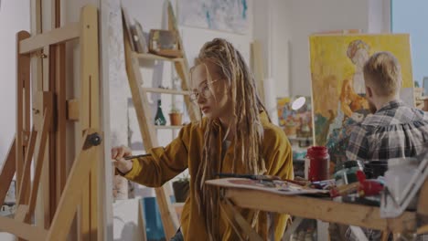 Pretty-Stylsih-Hipster-Girl-Artist-With-Dreadlocks-And-In-Glasses-Drawing-On-Canvas-With-Oil-Paints