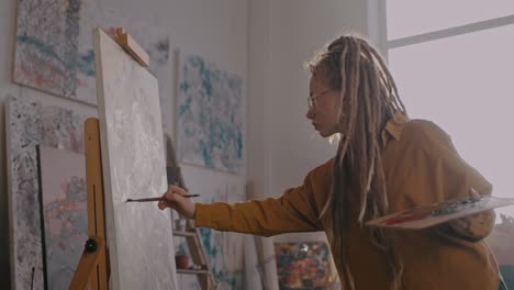Young-Female-Painter-With-Dreadlocks-And-In-Glasses-Painting-On-Canvas-At-Easel-With-Paints-And-Brush