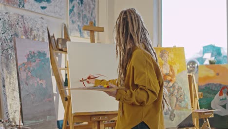 Back-View-On-Young-Female-Painter-With-Dreadlocks-Painting-On-Canvas-At-Easel-With-Red-Paint-And-Brush