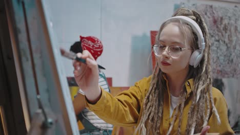 Close-Up-Of-Young-â€“Â°Auc-Beautiful-Girl-In-Glasses-And-Dreadlocks-With-Headphones-Drawing-At-Easel-In-Studio