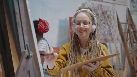 Portrait-Of-Young-Stylish-Female-Artist-In-Glasses,-Headphones-And-With-Dreadlocks-Painting-Picture-On-Canvas-And-Listening-To-Music