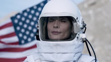 Portrait-Of-A-Female-Astronaut-Wearing-A-Helmet-Looking-Straight-Ahead-With-An-American-Flag-In-The-Background