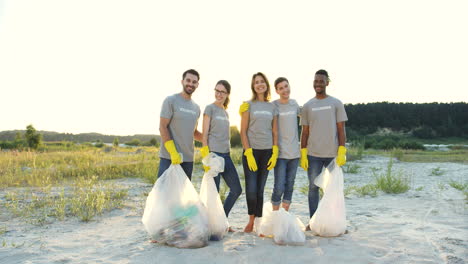 Portrait-Shot-Of-The-Mixed-Races-People-Standing-In-Front-Of-The-Camera-With-Trash-Bags-After-Collecting-Garbage-Near-Lake-Or-River
