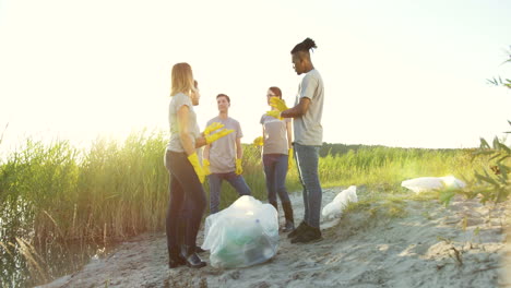 Young-Multiethnical-Volunteers-Standing-At-The-Lake-Or-Sea-Beach-And-Talking-While-Collecting-Litter-In-The-Plastic-Bags