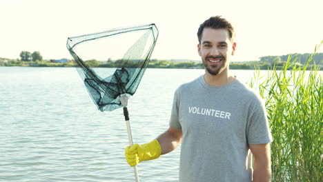 Young-Handsome-Man-Smiling-And-Posing-To-The-Camera-With-A-Steel-Net-Full-Of-Garbage-After-Cleaning-The-Lake-Bottom